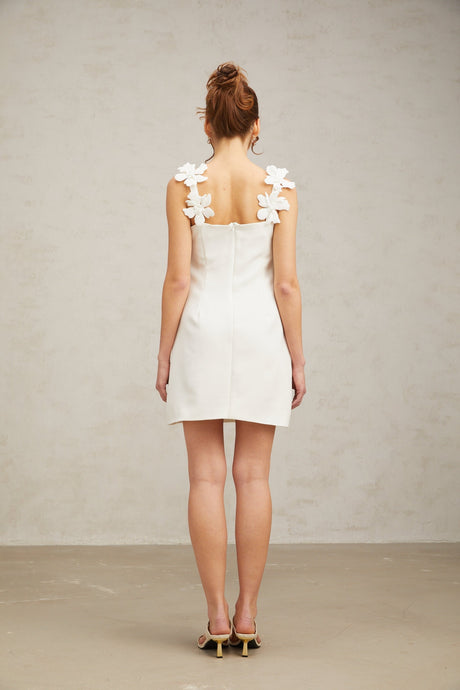 Young woman wearing a short white dress with floral embroidery standing in front of a plain background