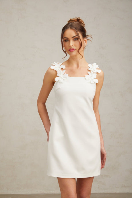 Young woman wearing a short white dress with floral lace pattern and ruffle details standing in front of a plain background