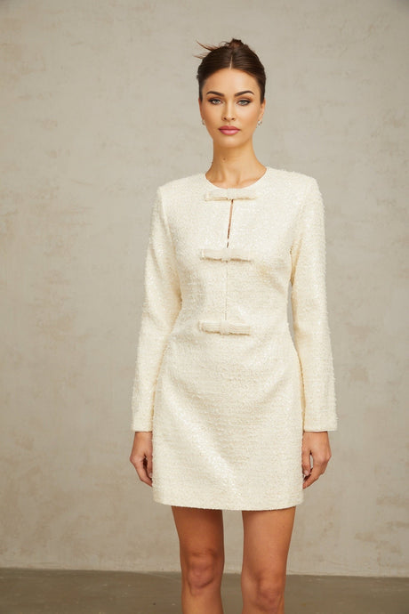 Young woman wearing a short white dress with sequin embellishments and tweed texture standing against a plain background