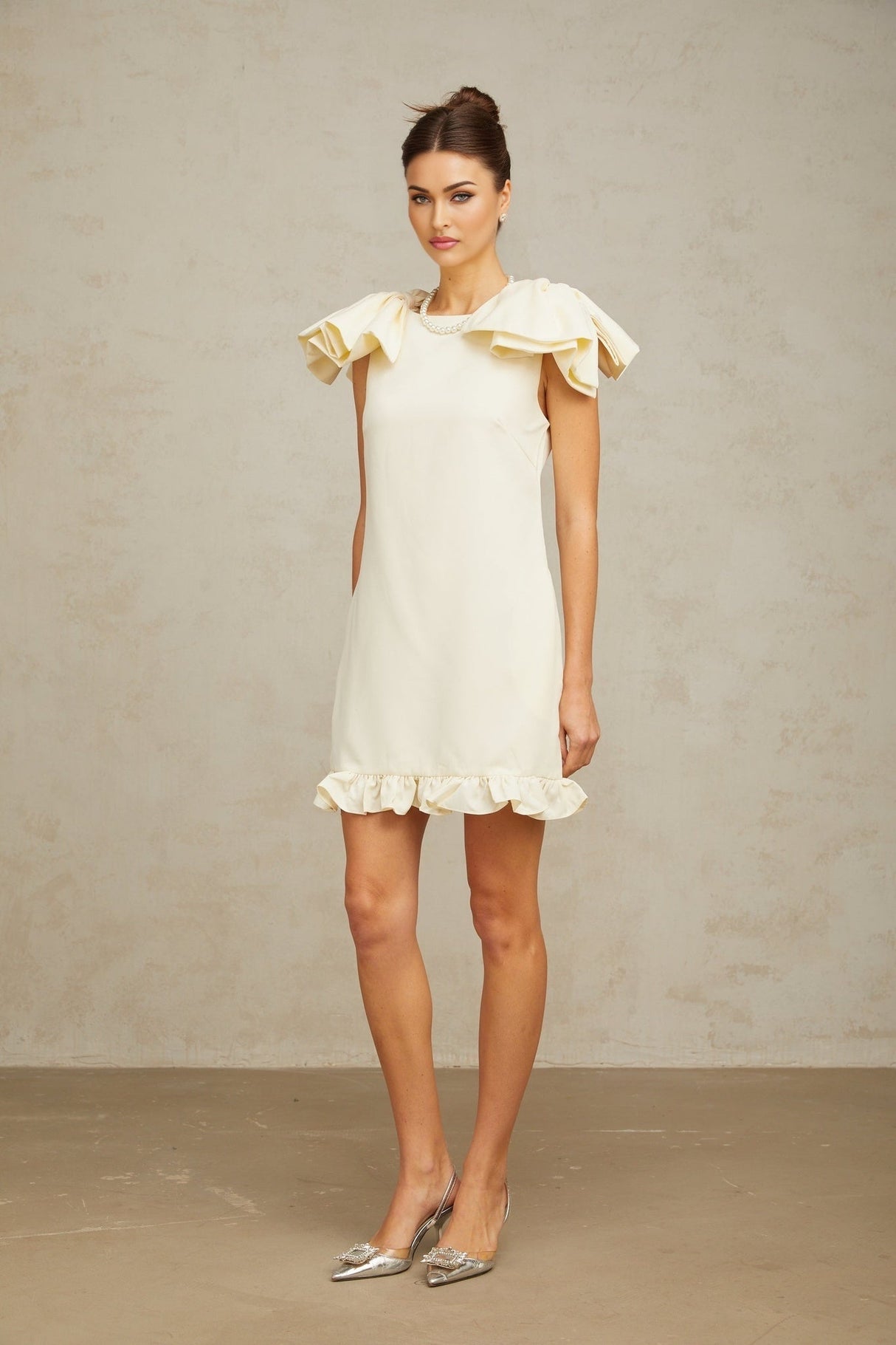 Young woman wearing a short white mini dress with ruffled details and a bow accent standing against a plain background