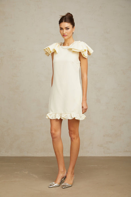Young woman wearing a short white mini dress with ruffled details and a bow accent standing against a plain background