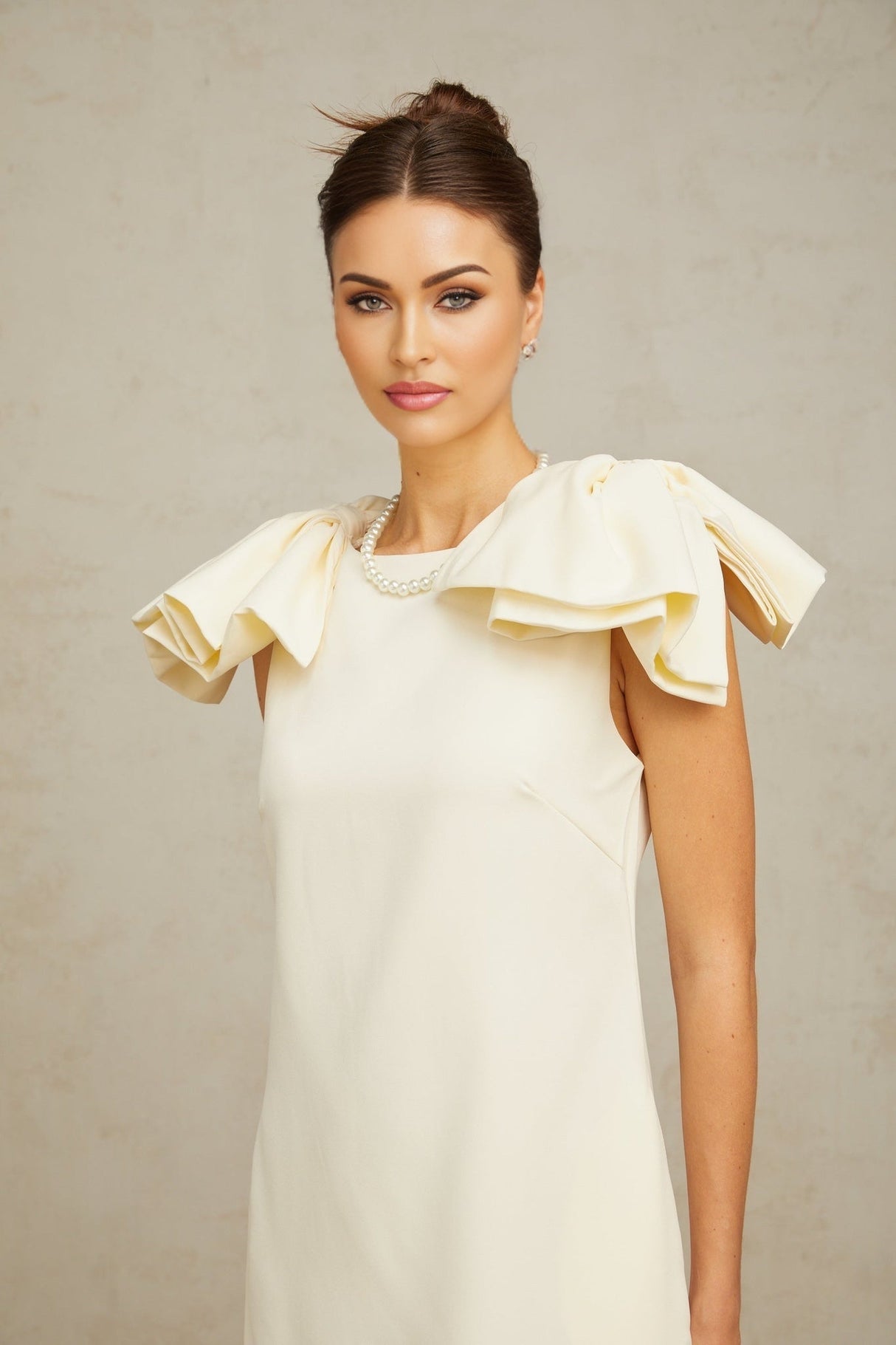 Young woman wearing a short white mini dress with ruffled details and a bow accent standing against a plain background