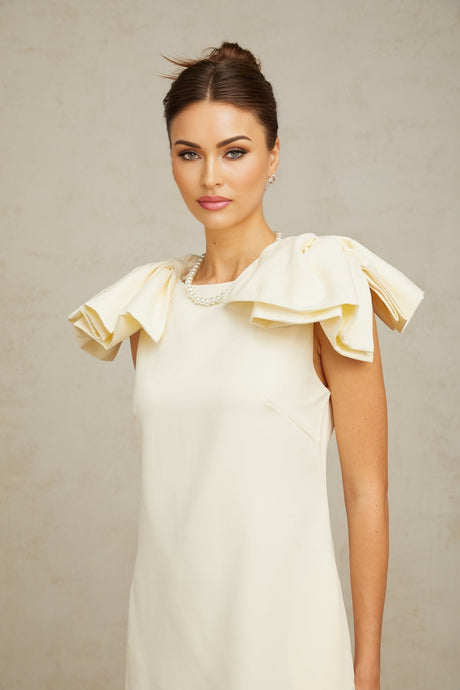 Young woman wearing a short white mini dress with ruffled details and a bow accent standing against a plain background