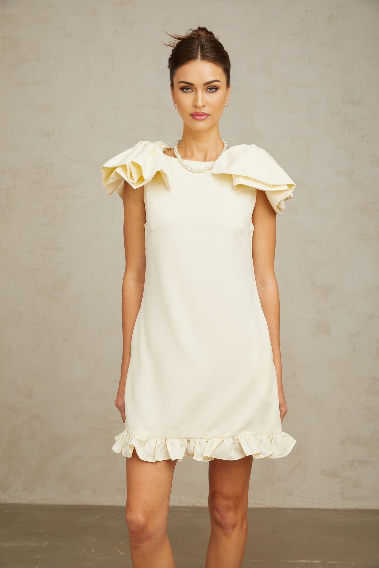 Young woman wearing a short white mini dress with ruffled details and a large bow accent standing against a plain background