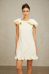 Young woman wearing a short white mini dress with ruffled details and a large bow accent standing against a plain background