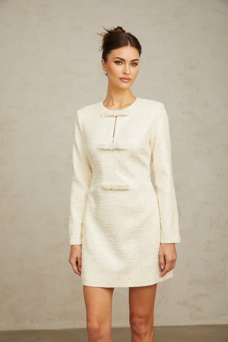 Young woman wearing a short white tweed dress with sequin embellishments standing against a plain background