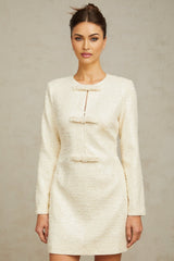 Young woman wearing a short white tweed dress with sequin embellishments standing in front of a plain background