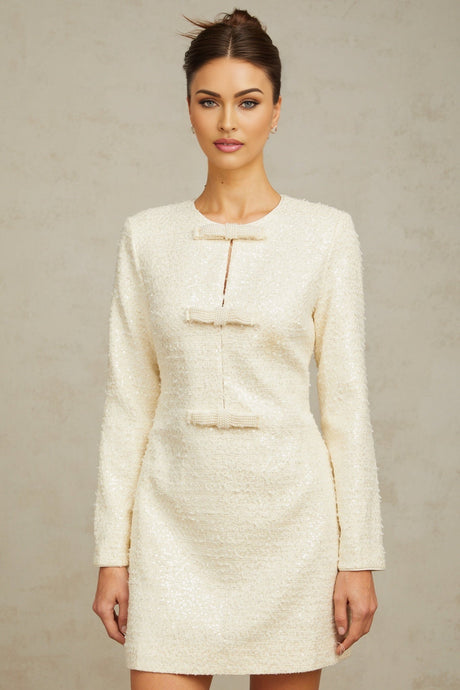 Young woman wearing a short white tweed dress with sequin embellishments standing in front of a plain background