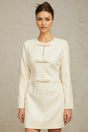 Young woman wearing a short white tweed dress with sequin embellishments standing in front of a plain background