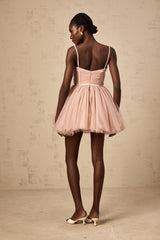 Young woman wearing a sleeveless pleated tulle mini dress in a light color standing against a plain background