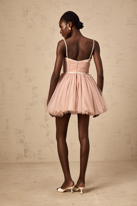Young woman wearing a sleeveless pleated tulle mini dress in a light color standing against a plain background