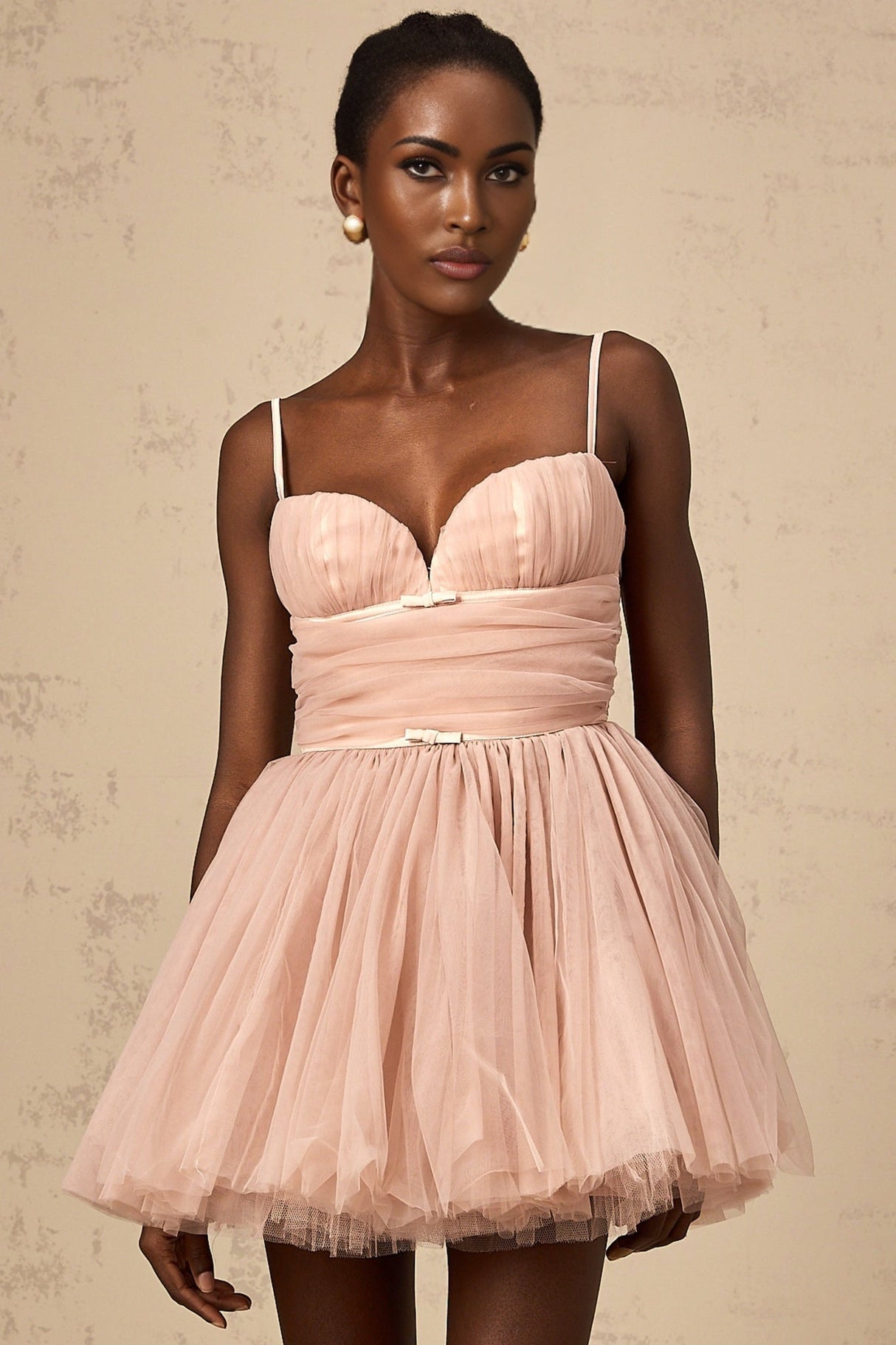 Young woman wearing a sleeveless pleated tulle mini dress in a light color standing against a plain background