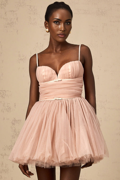 Young woman wearing a sleeveless pleated tulle mini dress in a light color standing against a plain background
