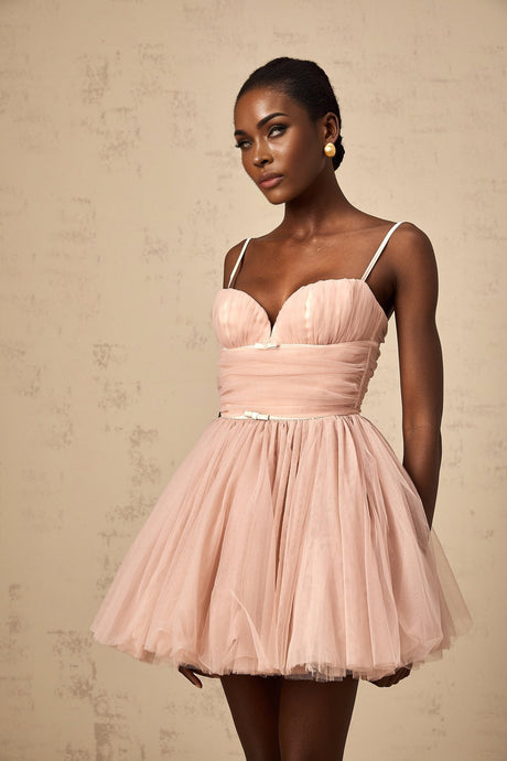 Young woman wearing a sleeveless pleated tulle mini dress in a light color standing against a plain background