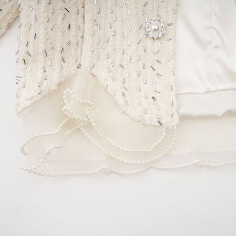 Young woman wearing a white cropped jacket with beaded details and ruffle trim standing against a plain background