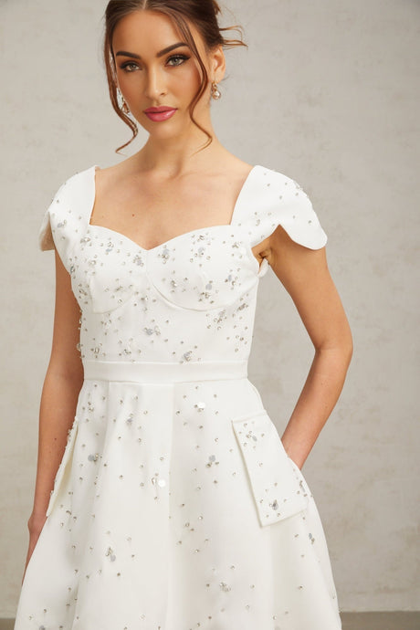 Young woman wearing a white flared mini dress with embellishments standing against a plain background