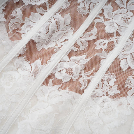 Young woman wearing a white floral lace corset mini dress with delicate straps and a fitted bodice standing against a plain background