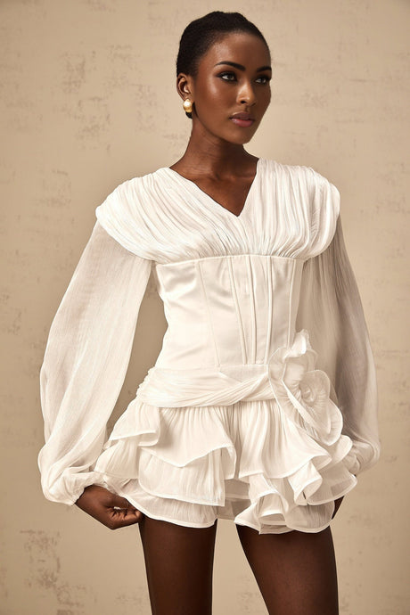 Young woman wearing a white fully pleated mini dress with cinched waist standing against plain background