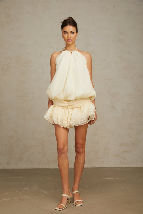 Young woman wearing a white halterneck mini dress adorned with faux pearls standing against a plain background
