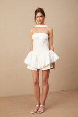 Young woman wearing a white halterneck mini dress with ruffle details standing against a plain background