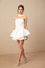 Young woman wearing a white halterneck mini dress with ruffled details standing against a plain background