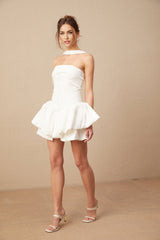 Young woman wearing a white halterneck mini dress with ruffled details standing against a plain background