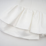 Young woman wearing a white halterneck mini dress with ruffled details standing against a plain background