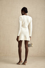 Young woman wearing a white knitted mini dress with flared skirt standing against a plain background