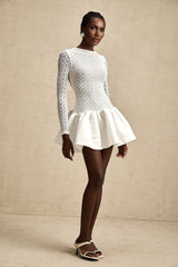 Young woman wearing a white lace mini dress with panel details standing against a plain background