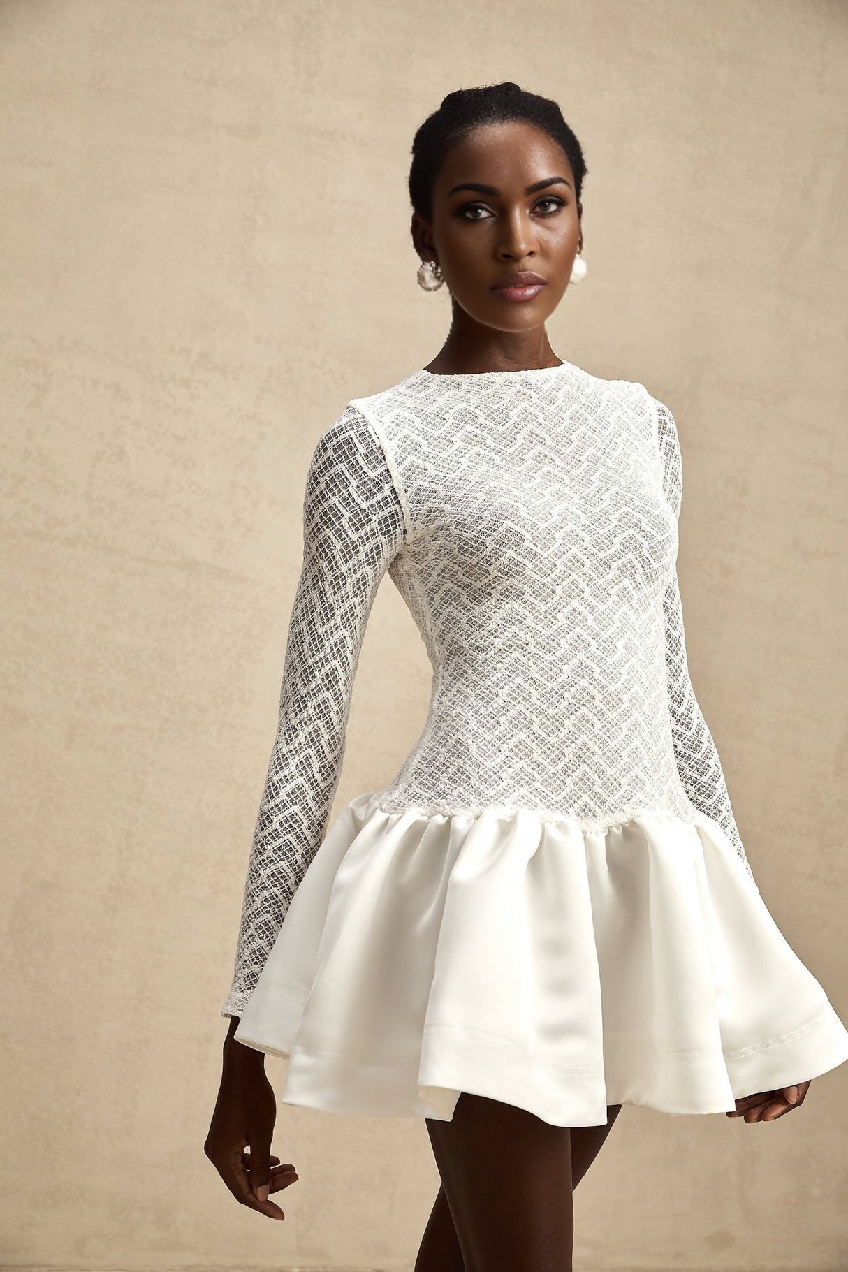 Young woman wearing a white lace mini dress with panelled design standing against a plain background