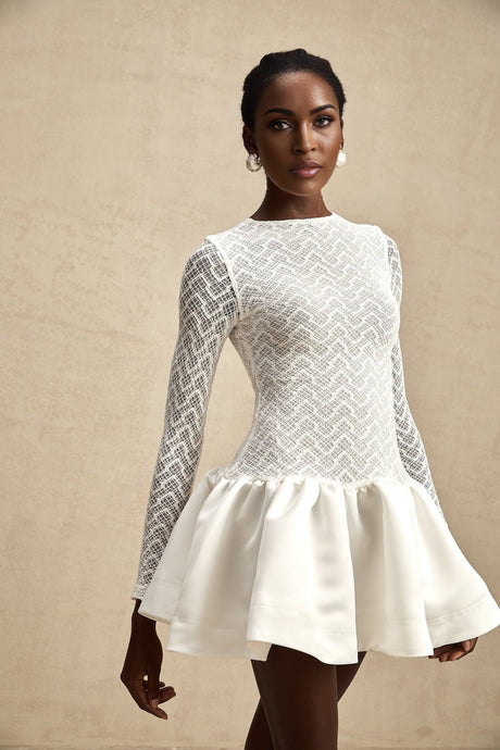 Young woman wearing a white lace mini dress with panelled design standing against a plain background
