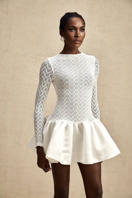 Young woman wearing a white lace mini dress with sheer panels standing against a plain background