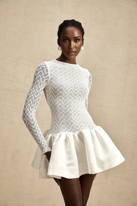 Young woman wearing a white lace mini dress with sheer panels standing in front of a plain background