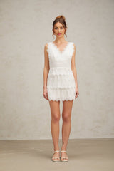 Young woman wearing a white lace mini dress with tiered skirt and short sleeves standing against a plain background