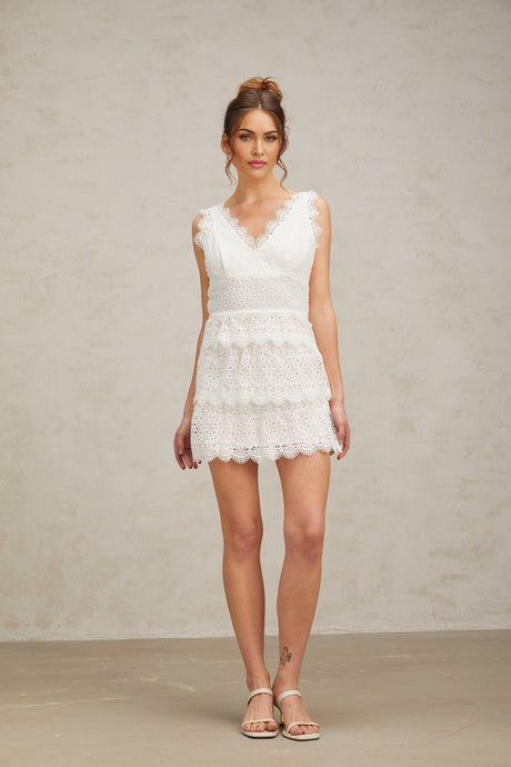 Young woman wearing a white lace tiered mini dress with short sleeves standing against a plain background