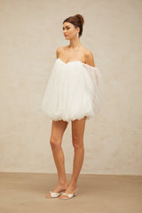 Young woman wearing a white layered tulle mini dress with bubble sleeves and a flared skirt standing against a plain background