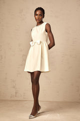 Young woman wearing a white mini dress with a bowknot embellishment standing against a plain background