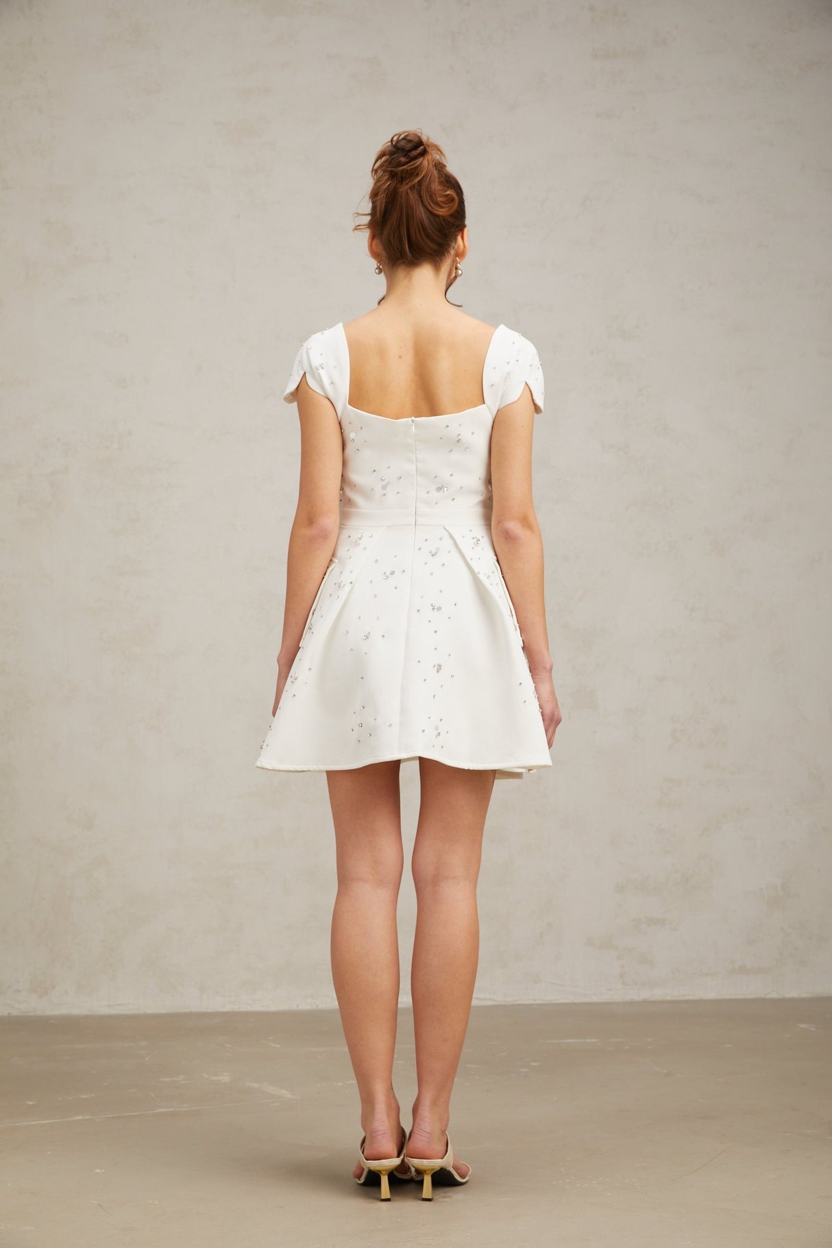 Young woman wearing a white mini dress with embellishments and a flared skirt standing against a plain background