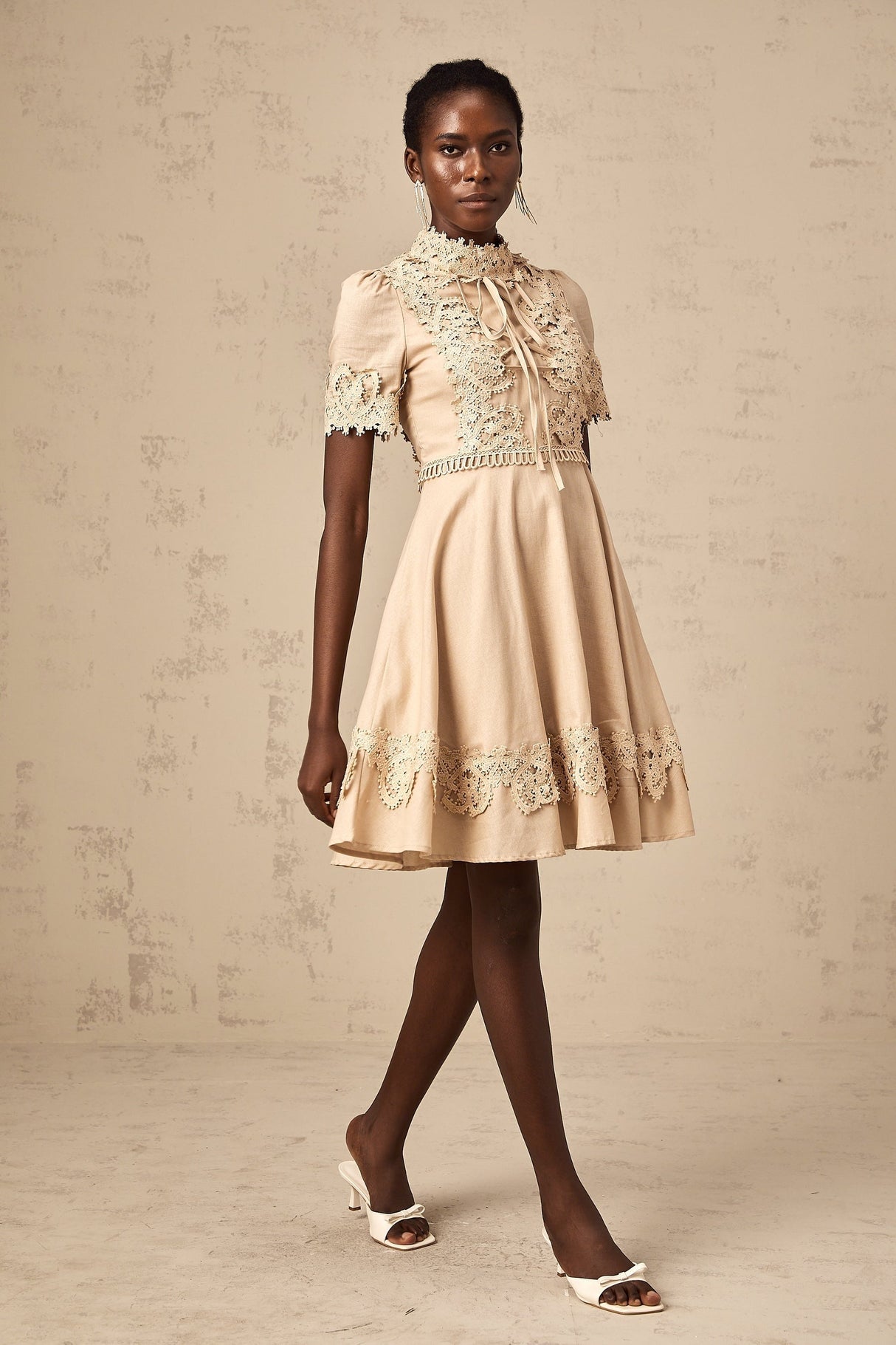 Young woman wearing a white mini dress with flared skirt and corded lace details standing in front of neutral background