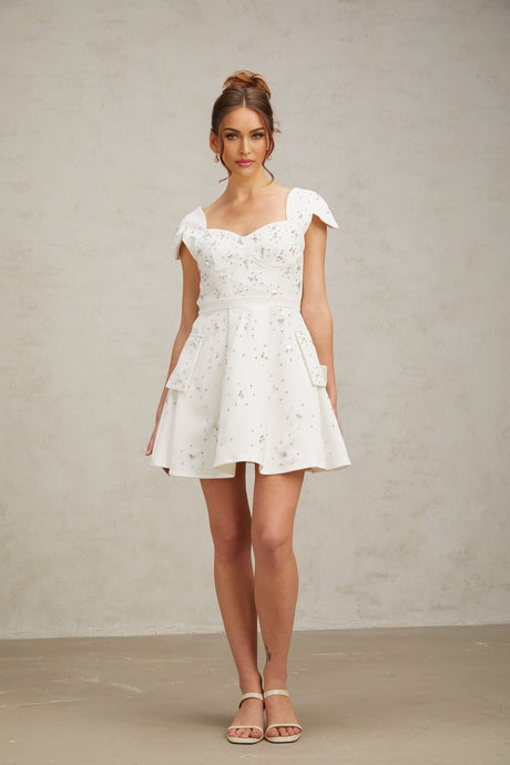 Young woman wearing a white mini dress with flared skirt and embellished details standing against a plain background