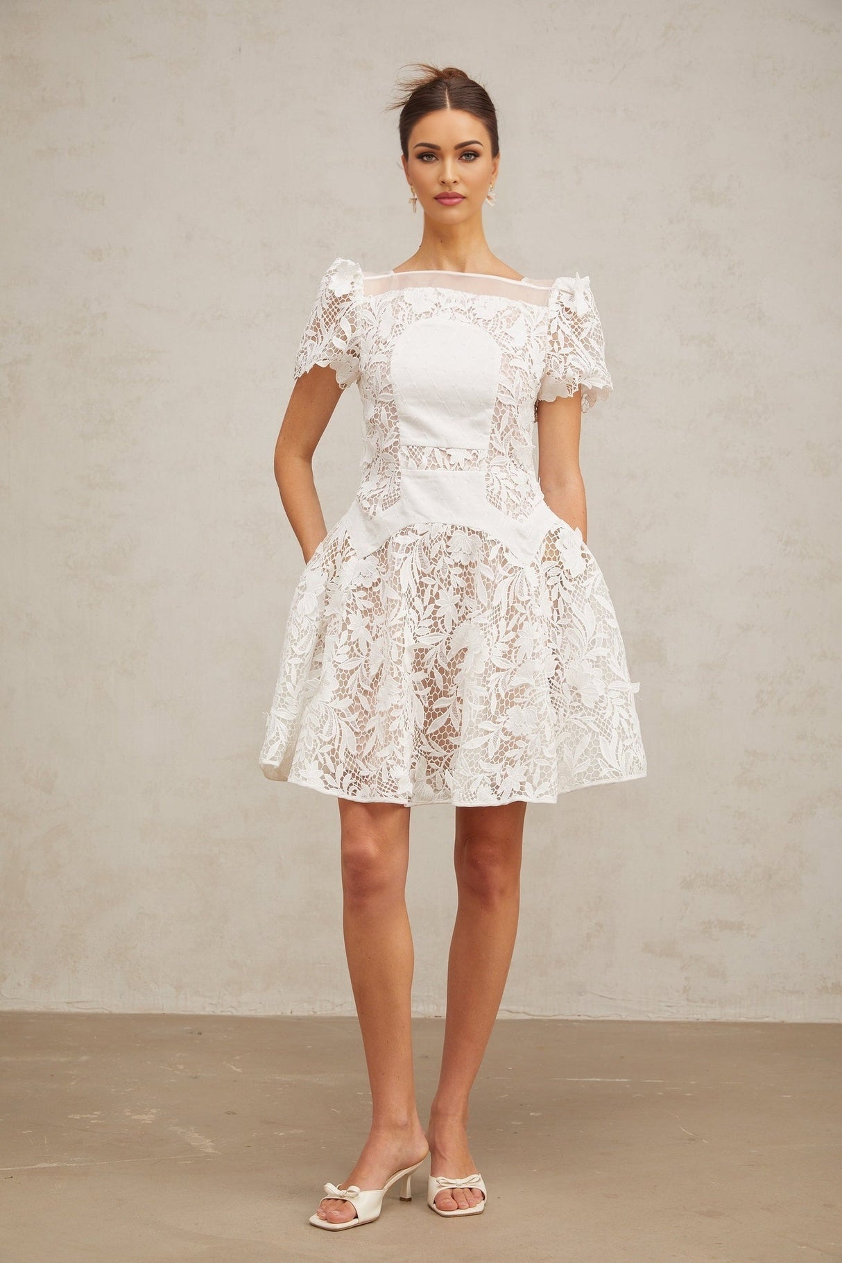 Young woman wearing a white mini dress with floral lace pattern and puff sleeves standing against a plain background