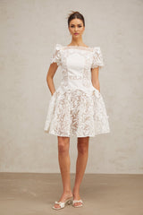 Young woman wearing a white mini dress with floral lace pattern and puff sleeves standing against a plain background