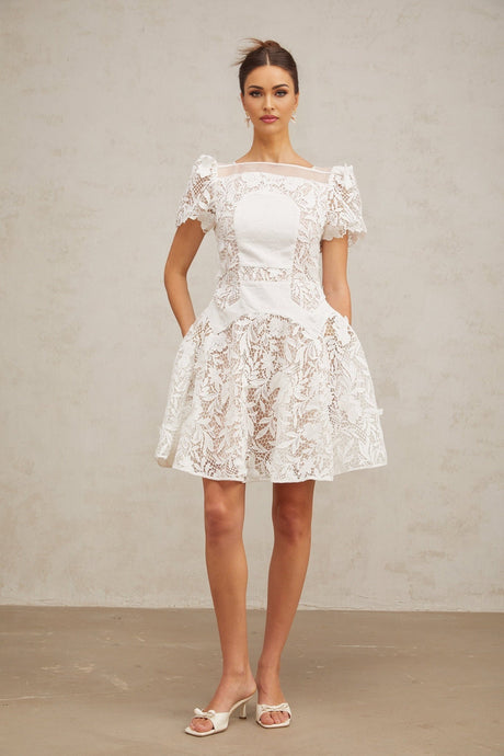 Young woman wearing a white mini dress with floral lace pattern and puff sleeves standing against a plain background