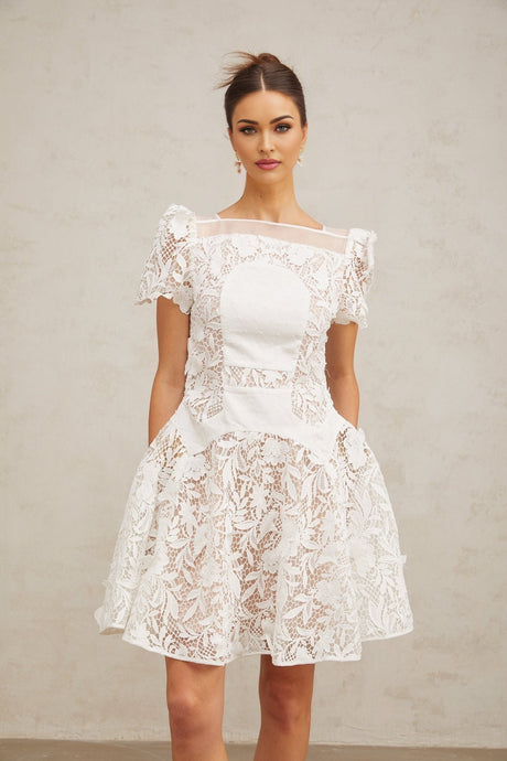 Young woman wearing a white mini dress with floral lace pattern and puff sleeves standing against a plain background