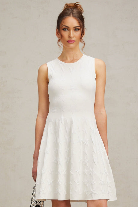 Young woman wearing a white mini dress with jasmine star pattern knitted design standing against plain background