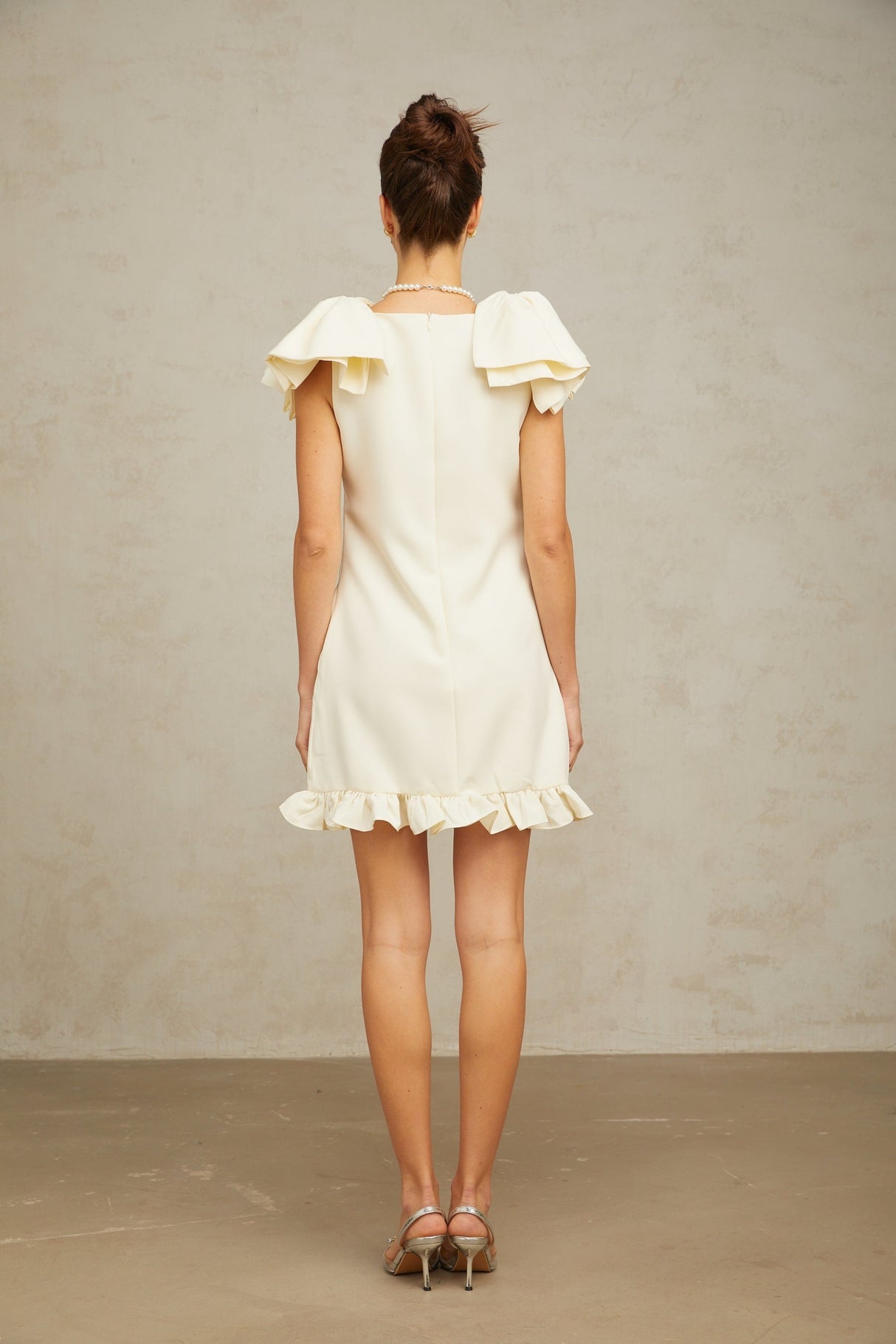 Young woman wearing a white mini dress with ruffled details and a bow accent standing against a plain background