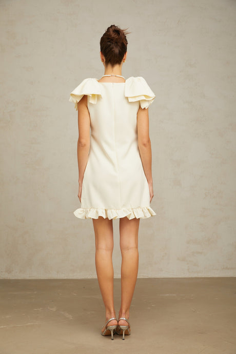 Young woman wearing a white mini dress with ruffled details and a bow accent standing against a plain background