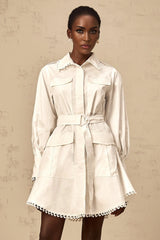 Young woman wearing a white mini shirt dress with a belt cinched at the waist standing against a plain background