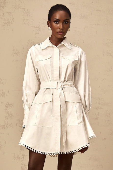 Young woman wearing a white mini shirt dress with a belt cinched at the waist standing against a plain background
