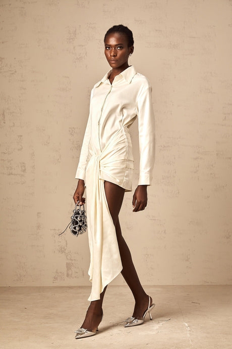Young woman wearing a white mini shirt dress with ruched detailing and ribbon accents standing against a plain background